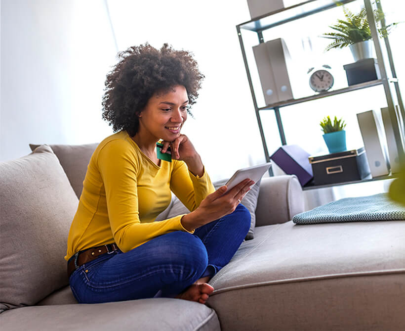 Woman sitting on a sofa looking at her tablet. Various household items in the background